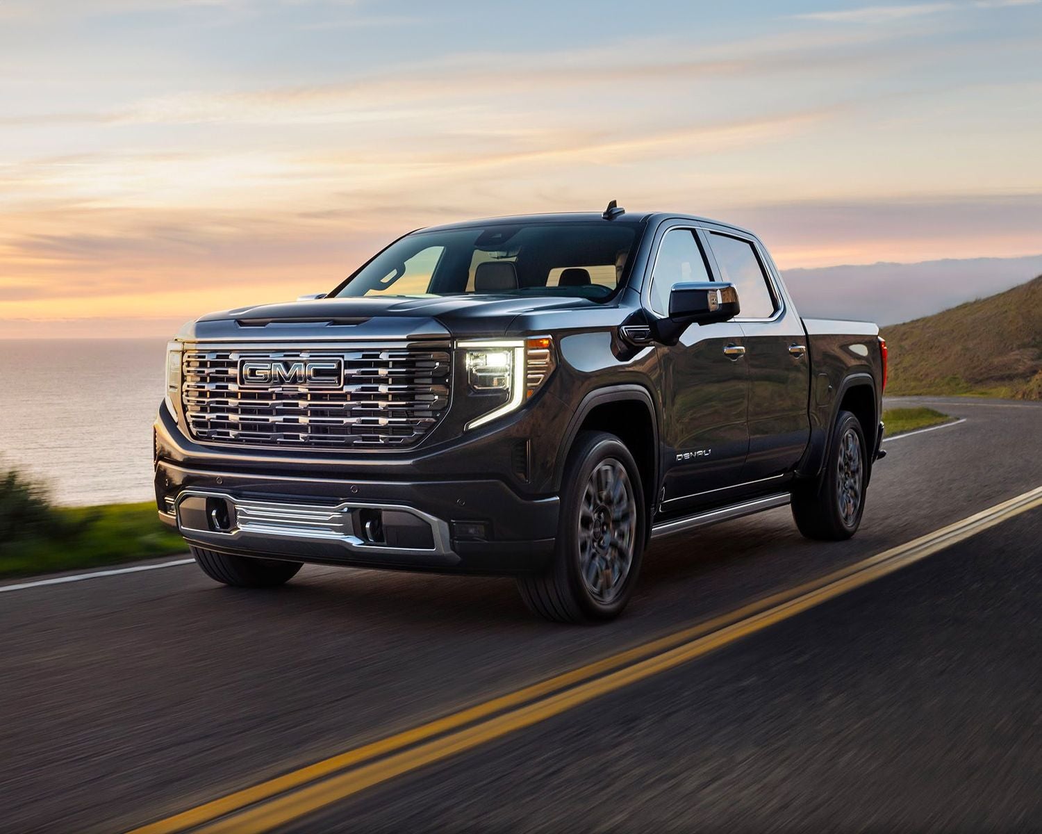 Front 3/4 shot of the 2024 GMC Sierra 1500 driving down mountain road at dusk with beautiful skyline.